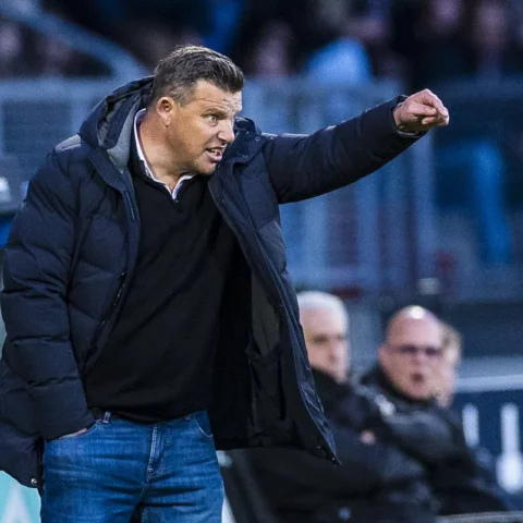 Een voetbaltrainer in een donkere jas wijst vanaf de zijlijn tijdens een wedstrijd.