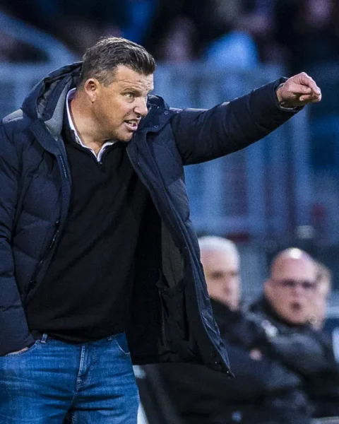 Een voetbaltrainer in een donkere jas wijst vanaf de zijlijn tijdens een wedstrijd.
