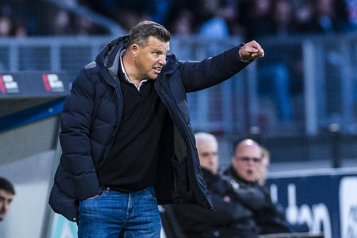 Een voetbaltrainer in een donkere jas wijst vanaf de zijlijn tijdens een wedstrijd.