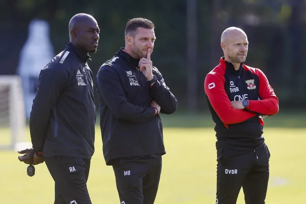 Drie voetbaltrainers staan langs het trainingsveld in clubkleding, met de middelste man denkend met een vinger tegen zijn lippen.