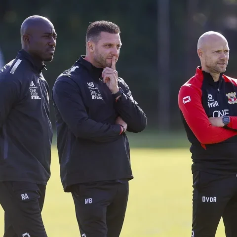 Drie voetbaltrainers staan langs het trainingsveld in clubkleding, met de middelste man denkend met een vinger tegen zijn lippen.