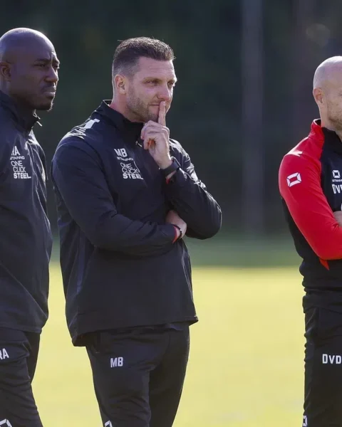 Drie voetbaltrainers staan langs het trainingsveld in clubkleding, met de middelste man denkend met een vinger tegen zijn lippen.