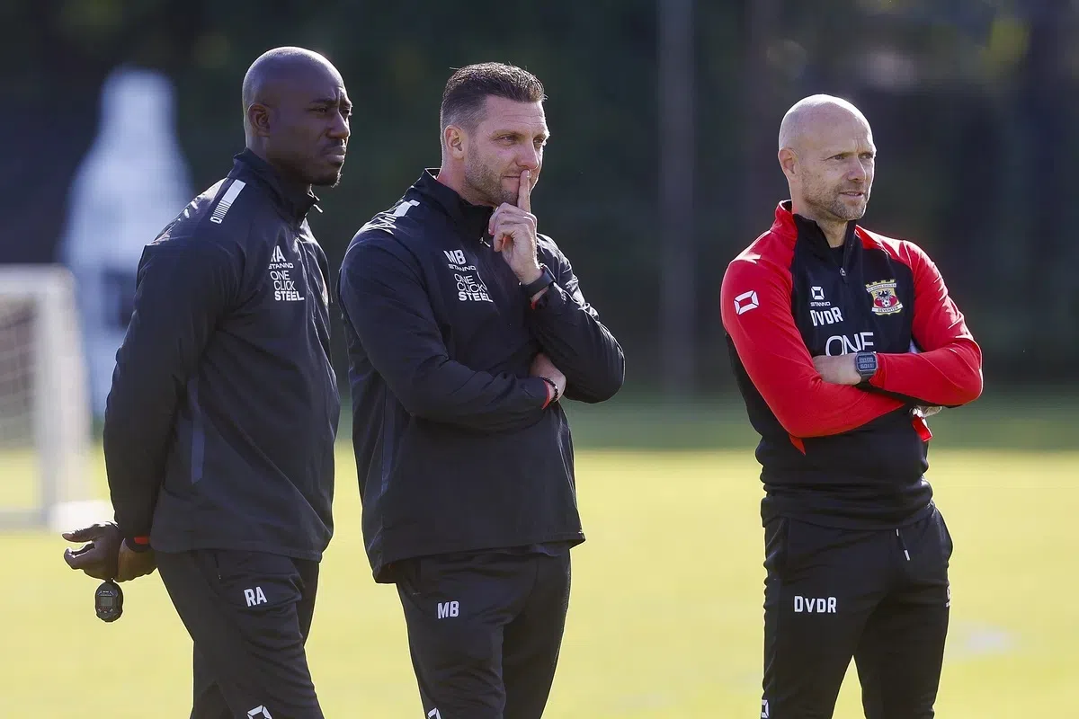 Drie voetbaltrainers staan langs het trainingsveld in clubkleding, met de middelste man denkend met een vinger tegen zijn lippen.