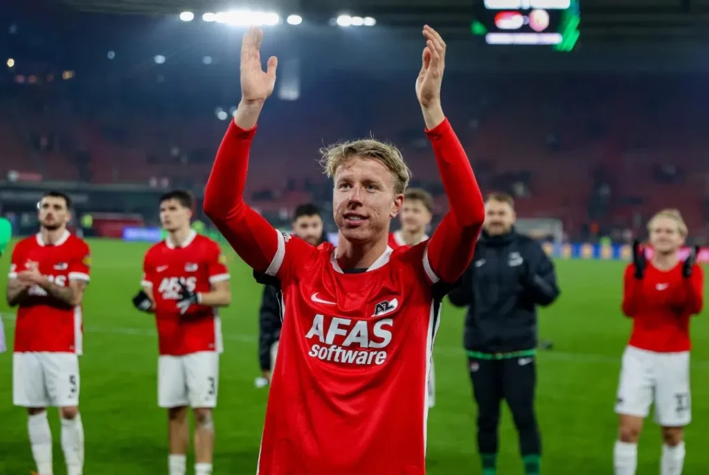 Billy van Duijl in een rood AZ-shirt op het veld met opgeheven armen, terwijl teamgenoten achter hem applaudisseren.