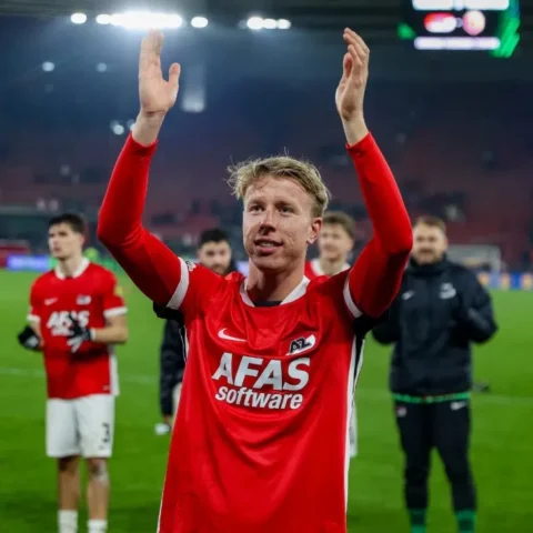 Billy van Duijl in een rood AZ-shirt op het veld met opgeheven armen, terwijl teamgenoten achter hem applaudisseren.