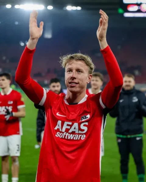 Billy van Duijl in een rood AZ-shirt op het veld met opgeheven armen, terwijl teamgenoten achter hem applaudisseren.
