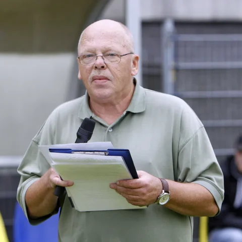 Een man met microfoon en klembord staat in een stadion.