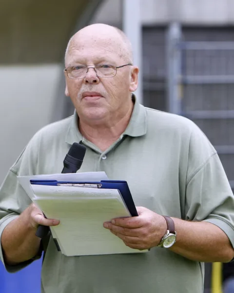 Een man met microfoon en klembord staat in een stadion.