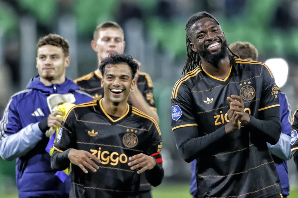 Ajax-spelers lachen en klappen op het veld, met Owen Wijndal niet zichtbaar in beeld.