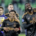 Ajax-spelers lachen en klappen op het veld, met Owen Wijndal niet zichtbaar in beeld.