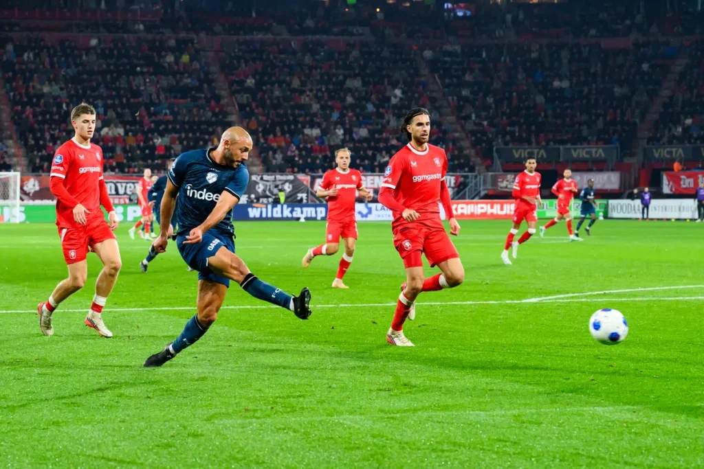 Voetballer schiet de bal tijdens een Eredivisie-wedstrijd tussen spelers in rode en donkerblauwe tenues.