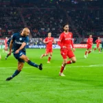 Voetballer schiet de bal tijdens een Eredivisie-wedstrijd tussen spelers in rode en donkerblauwe tenues.
