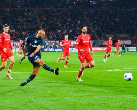 Voetballer schiet de bal tijdens een Eredivisie-wedstrijd tussen spelers in rode en donkerblauwe tenues.