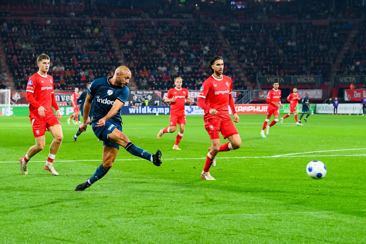 Voetballer schiet de bal tijdens een Eredivisie-wedstrijd tussen spelers in rode en donkerblauwe tenues.