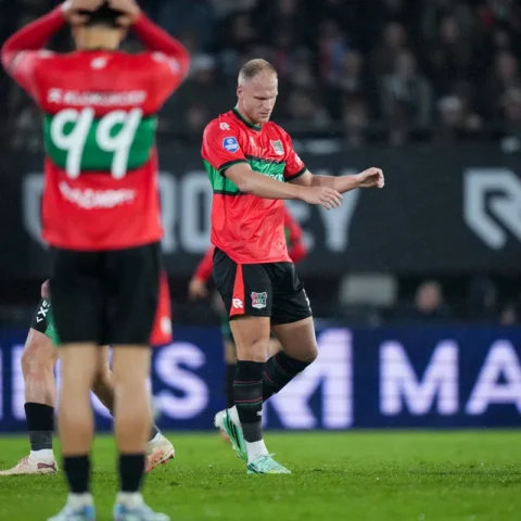 NEC-speler in rood-groen tenue op het veld tijdens een wedstrijd, met een teamgenoot op de voorgrond.