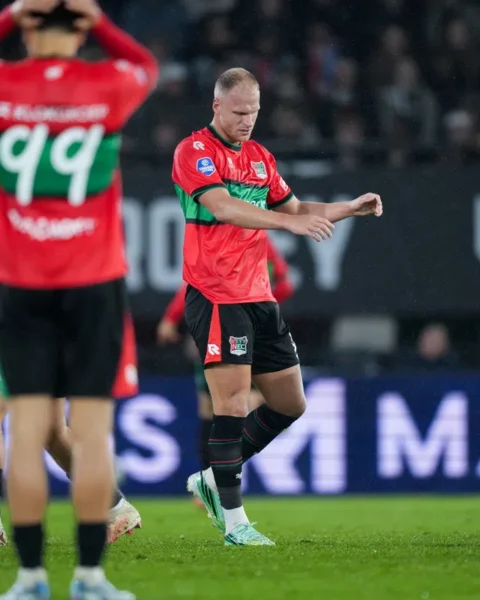 NEC-speler in rood-groen tenue op het veld tijdens een wedstrijd, met een teamgenoot op de voorgrond.