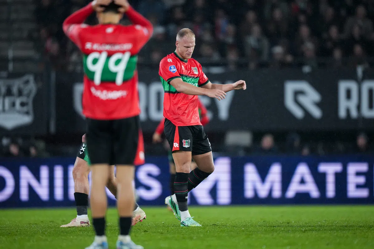 NEC-speler in rood-groen tenue op het veld tijdens een wedstrijd, met een teamgenoot op de voorgrond.