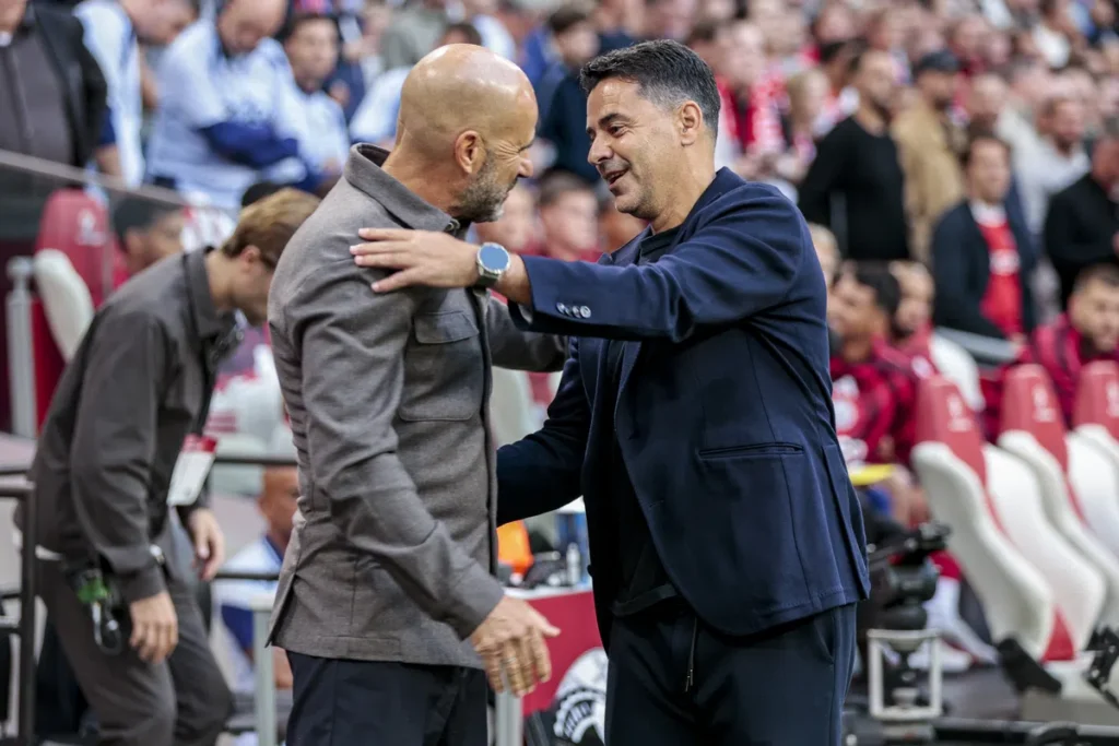 Twee voetbaltrainers begroeten elkaar langs het veld in een stadion.