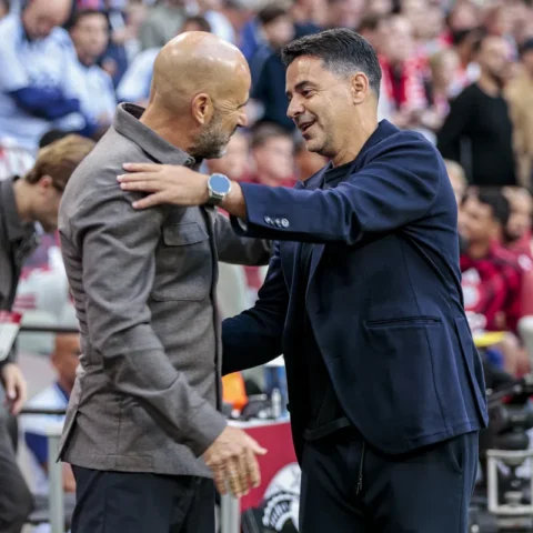 Twee voetbaltrainers begroeten elkaar langs het veld in een stadion.