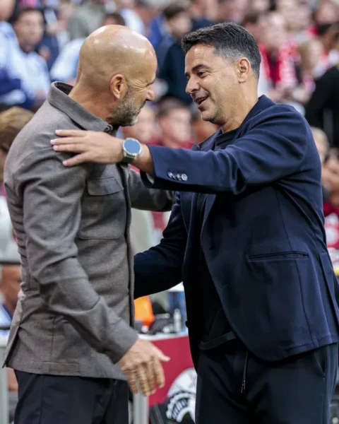 Twee voetbaltrainers begroeten elkaar langs het veld in een stadion.
