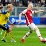 Twee voetballers in actie tijdens een wedstrijd, één in geel-blauw en één in rood-wit.