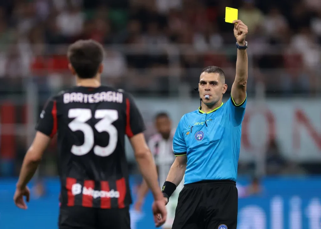 Scheidsrechter houdt een gele kaart omhoog tijdens een voetbalwedstrijd, terwijl een speler met rugnummer 33 toekijkt.