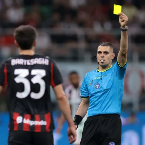 Scheidsrechter houdt een gele kaart omhoog tijdens een voetbalwedstrijd, terwijl een speler met rugnummer 33 toekijkt.