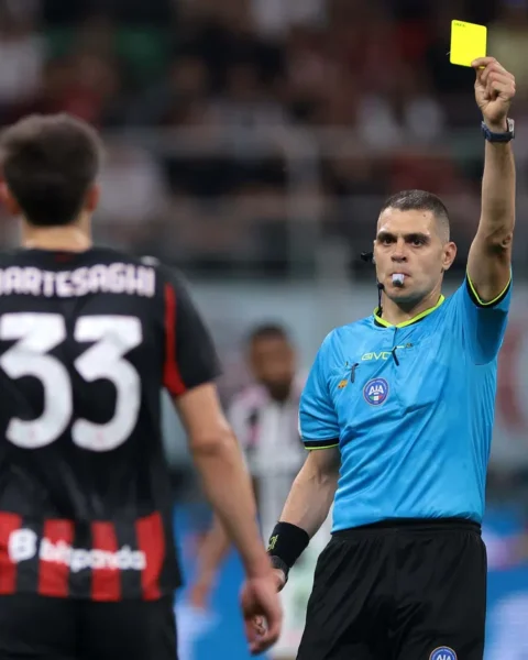 Scheidsrechter houdt een gele kaart omhoog tijdens een voetbalwedstrijd, terwijl een speler met rugnummer 33 toekijkt.