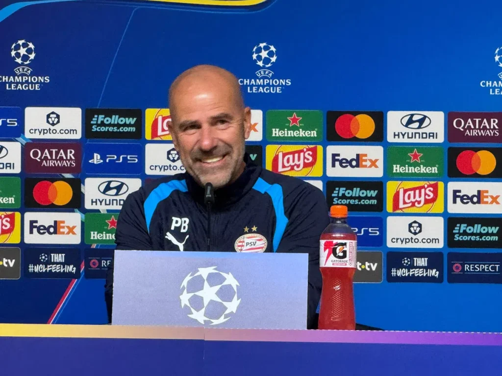 Peter Bosz zit achter een persbord bij een Champions League-persconferentie met PSV-logo en sponsorachtergrond.