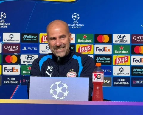 Peter Bosz zit achter een persbord bij een Champions League-persconferentie met PSV-logo en sponsorachtergrond.