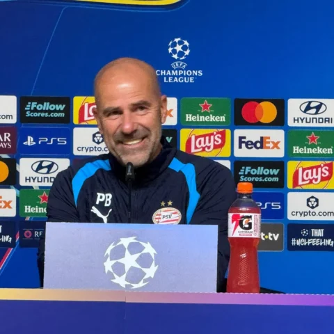 Peter Bosz zit achter een persbord bij een Champions League-persconferentie met PSV-logo en sponsorachtergrond.