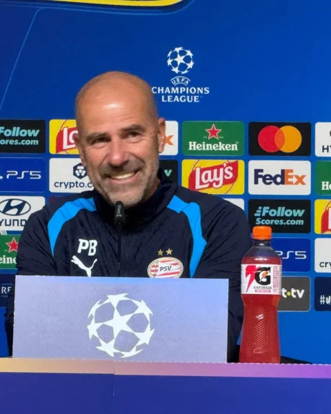Peter Bosz zit achter een persbord bij een Champions League-persconferentie met PSV-logo en sponsorachtergrond.