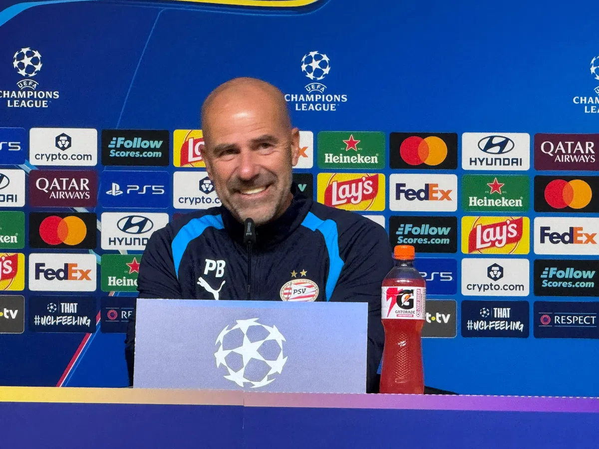 Peter Bosz zit achter een persbord bij een Champions League-persconferentie met PSV-logo en sponsorachtergrond.