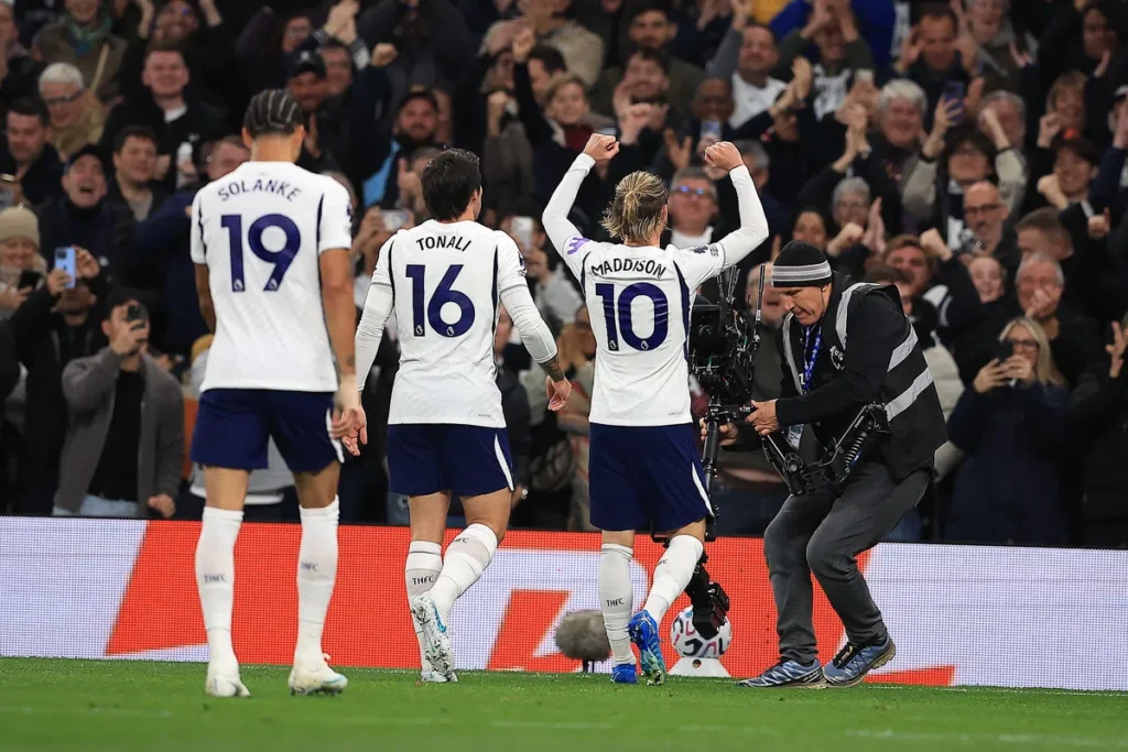 James Maddison viert een doelpunt voor Tottenham Hotspur, met teamgenoten naast hem en publiek op de achtergrond.