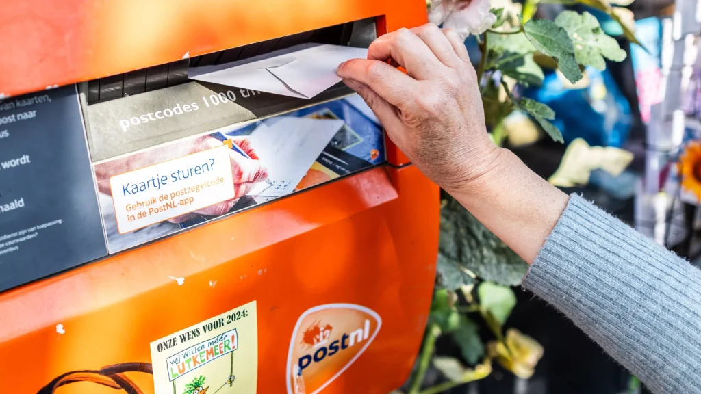Hand steekt een envelop in een oranje PostNL-brievenbus.