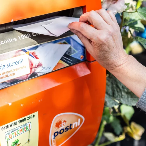 Hand steekt een envelop in een oranje PostNL-brievenbus.