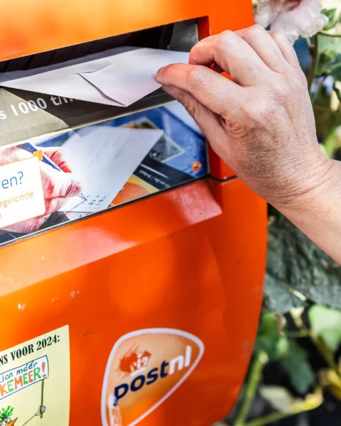 Hand steekt een envelop in een oranje PostNL-brievenbus.