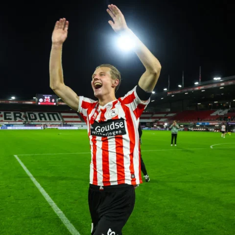 Een voetballer in rood-wit gestreept shirt juicht met beide armen omhoog op het veld.