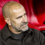 Peter Bosz zit op de bank in een rood stadionstoeltje.