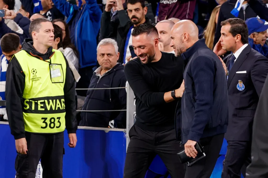 Peter Bosz lachend langs de zijlijn bij PSV, naast een andere man in donkere kleding, tijdens een Champions League-wedstrijd.