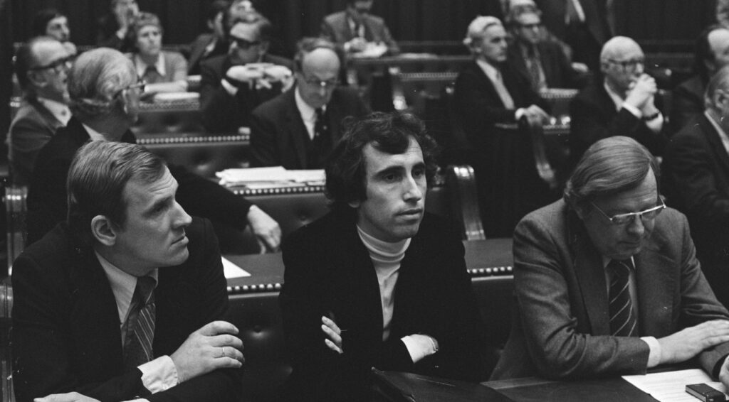 Zwart-witfoto van een parlementaire zaal met meerdere mannen in pakken op banken en stoelen.