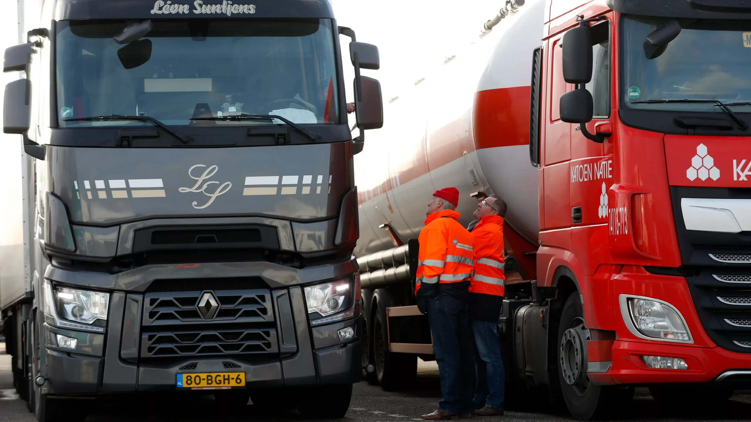 Twee vrachtwagens staan naast elkaar terwijl twee mannen in oranje veiligheidsjassen tussen de voertuigen staan.