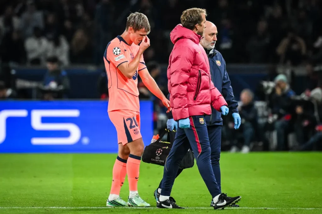 Dani Olmo staan op het veld terwijl hij zijn gezicht aanraakt, met Barcelona-staf naast hem.