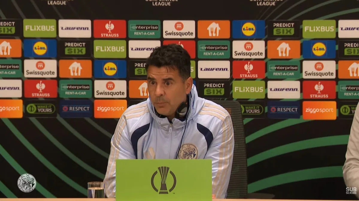 Trainer Míchel zit achter een tafel tijdens een persmoment met sponsorbord op de achtergrond.