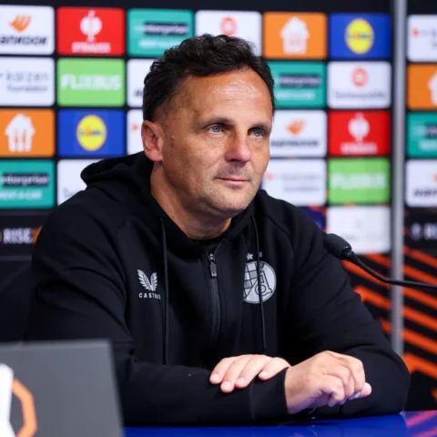 Een voetbaltrainer zit aan tafel tijdens een persconferentie voor een sponsorwand.