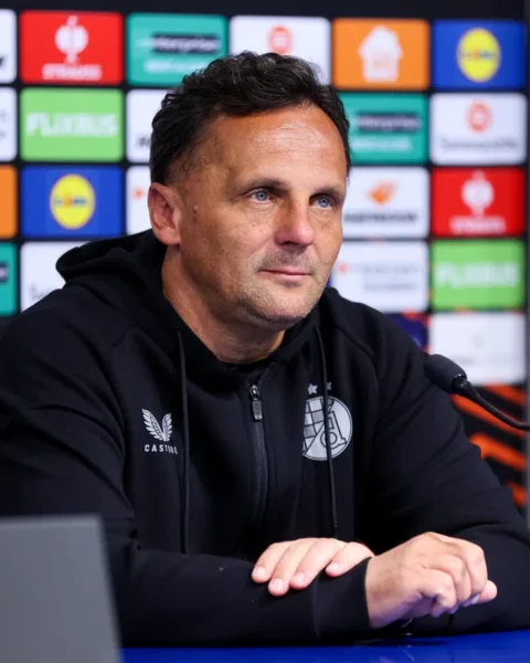 Een voetbaltrainer zit aan tafel tijdens een persconferentie voor een sponsorwand.