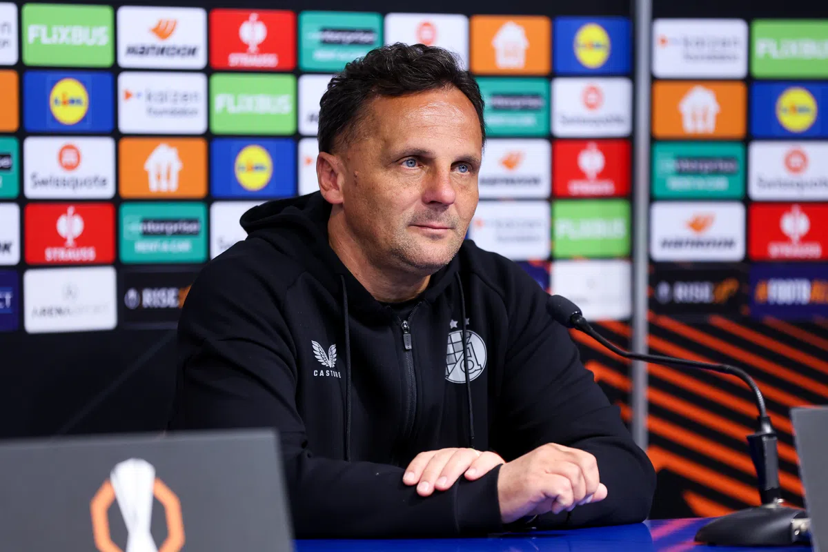 Een voetbaltrainer zit aan tafel tijdens een persconferentie voor een sponsorwand.