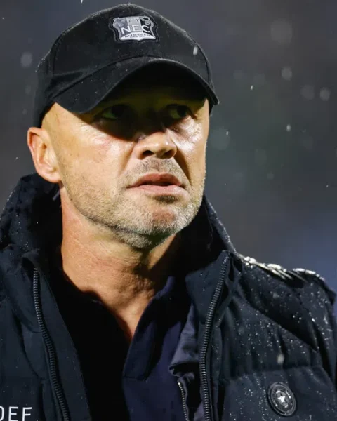 Dick Schreuder draagt een NEC-pet en zwarte jas in de regen.