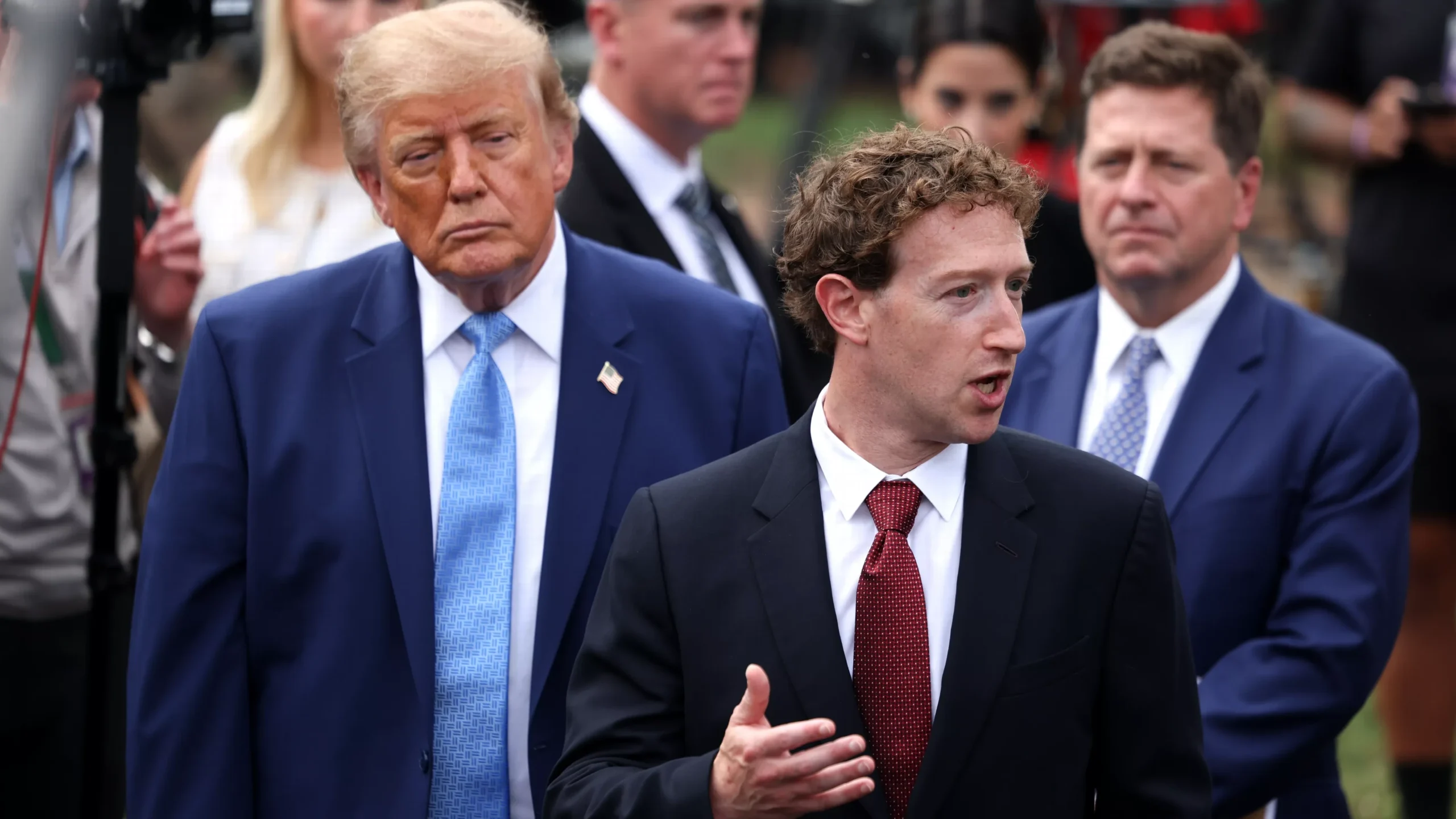 Mark Zuckerberg spreekt terwijl Donald Trump op de achtergrond staat, omringd door andere personen.