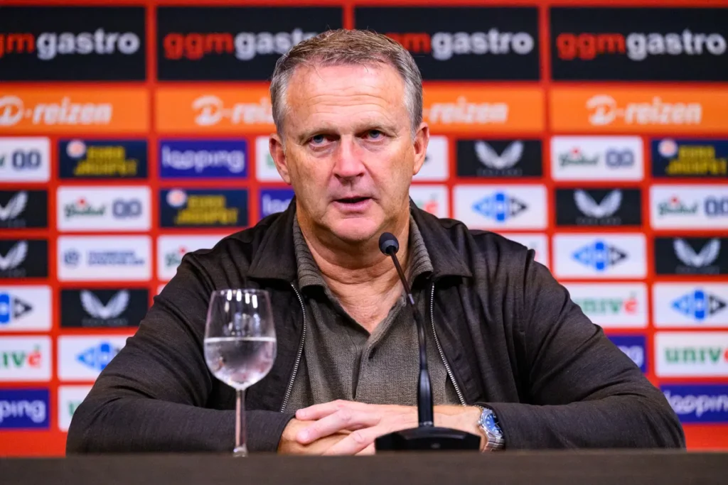 John van den Brom zit achter een microfoon tijdens een persmoment, met sponsorwand op de achtergrond.